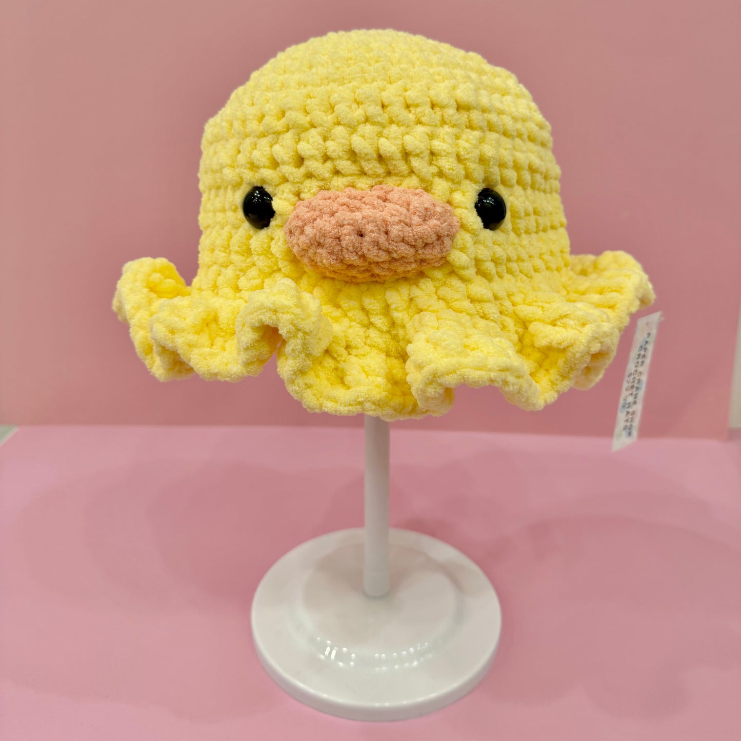 Yellow crochet chick hat on a white stand against a pink background