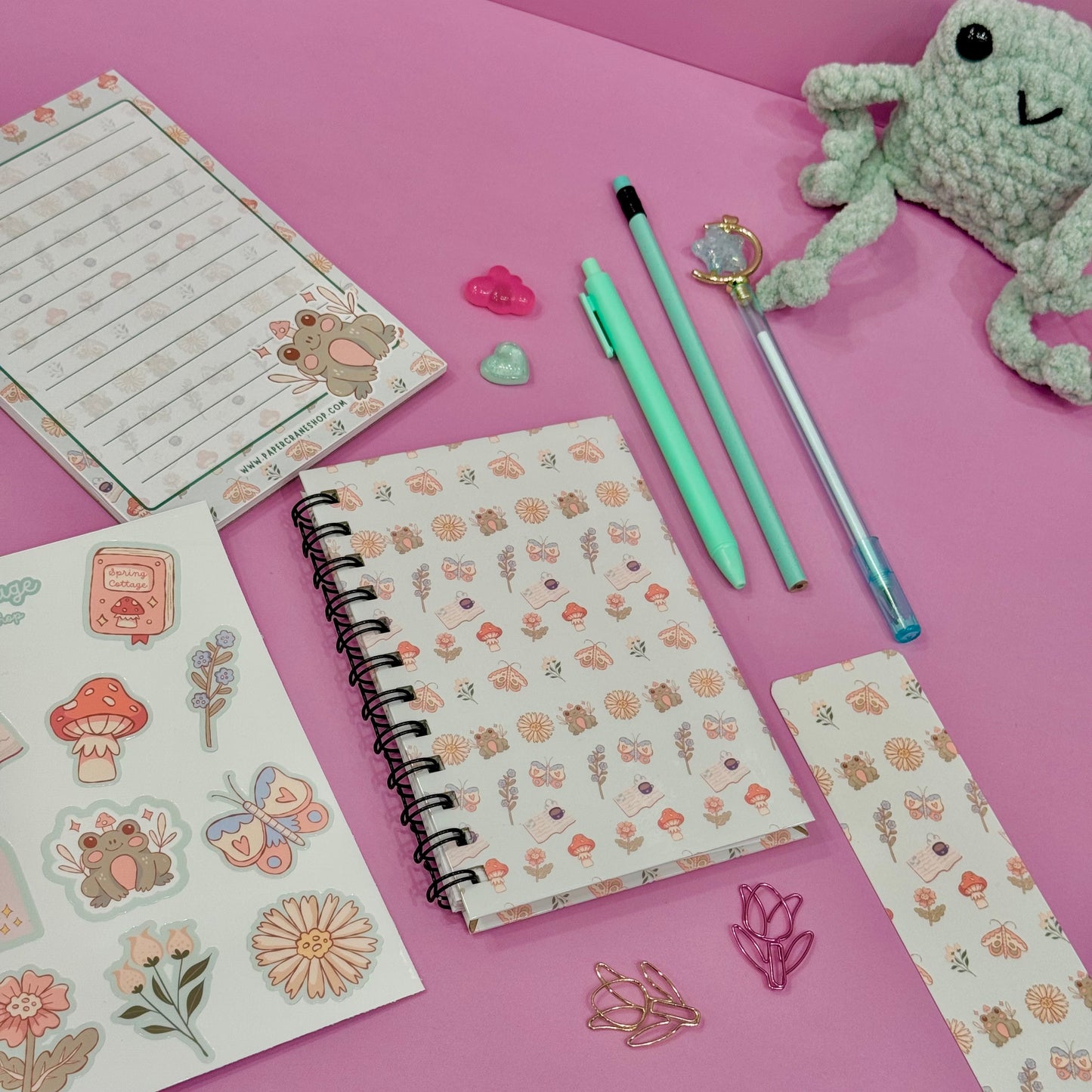 Stationery items including a notebook, pens, stickers, and a plush toy on a pink background