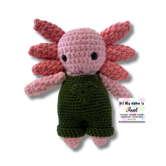 Crocheted axolotl toy with pink body and green overalls on a white background