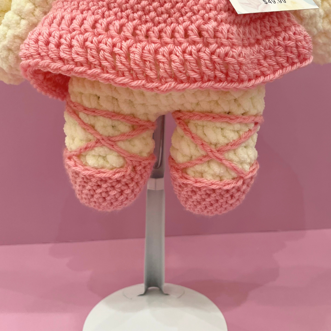 A close-up of a crocheted doll or toy torso and legs, with a pink textured top and cream-colored legs wrapped in pink yarn like ballet slippers, displayed on a stand against a pink background.