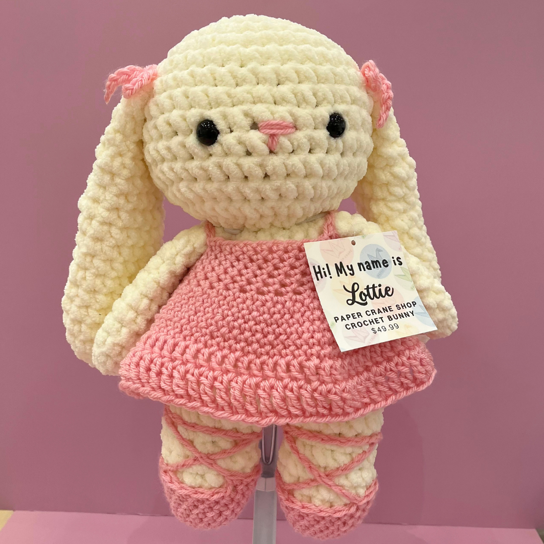 A handmade crocheted bunny doll named Littie, made with cream-colored yarn and a pink dress, displayed on a stand against a solid pink background. A paper tag on the dress reads 'Hi! My name is Lottie PAPER CANE SHOP CROCHET BUNNY 8.