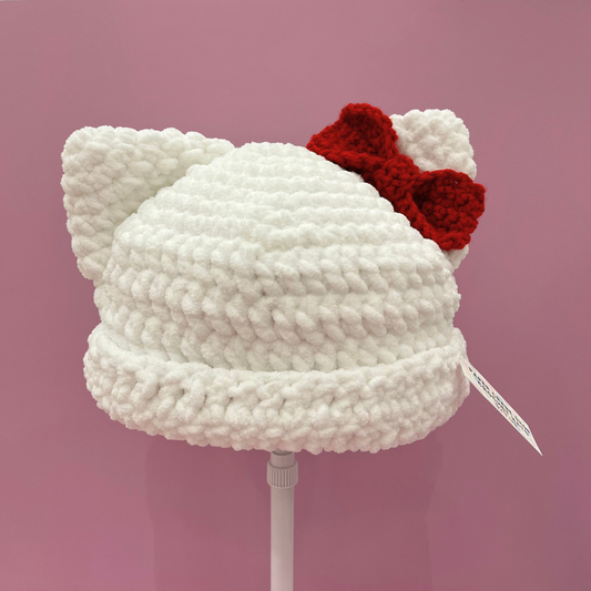 A white chunky crocheted cat-ear beanie hat with a red bow on top, displayed on a white stand against a solid pink background.