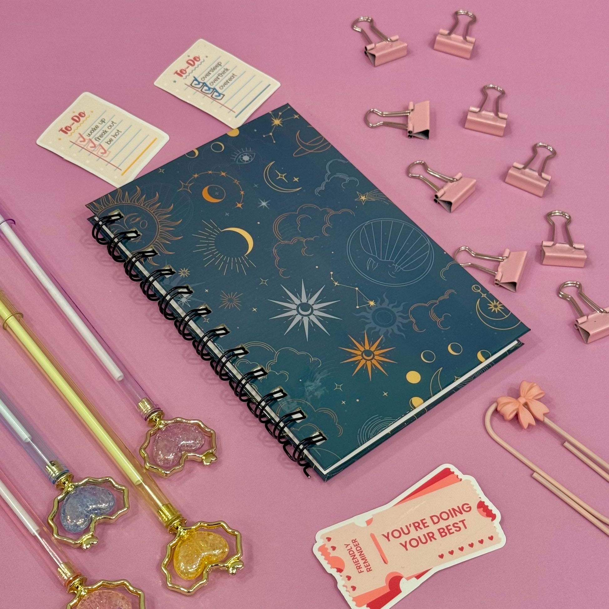 Notebook with celestial design surrounded by stationery items on a pink background