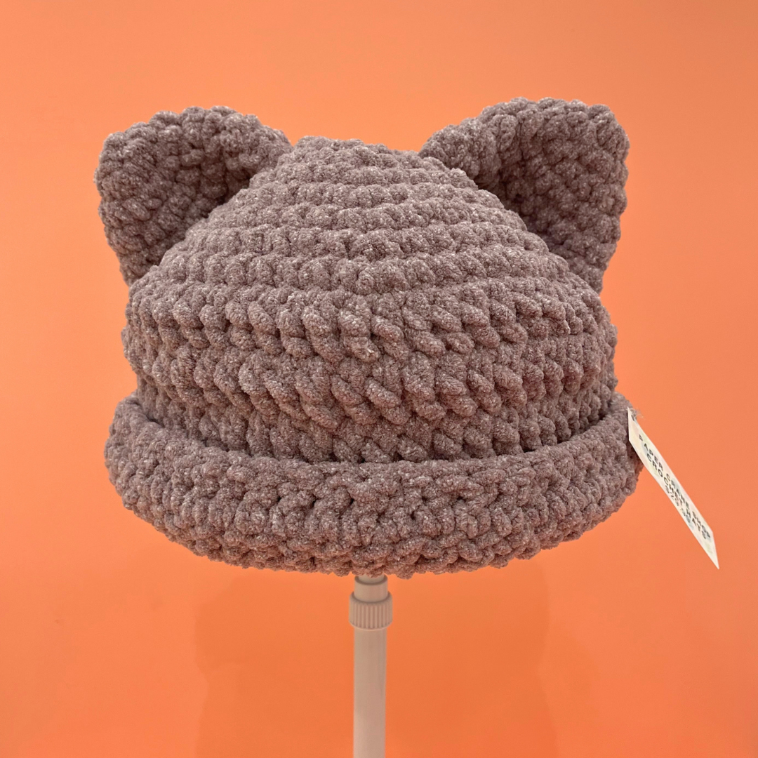 A chunky crocheted beanie hat with two small ears on top, shown on a stand against a solid orange background. The hat is a heathered purple color.