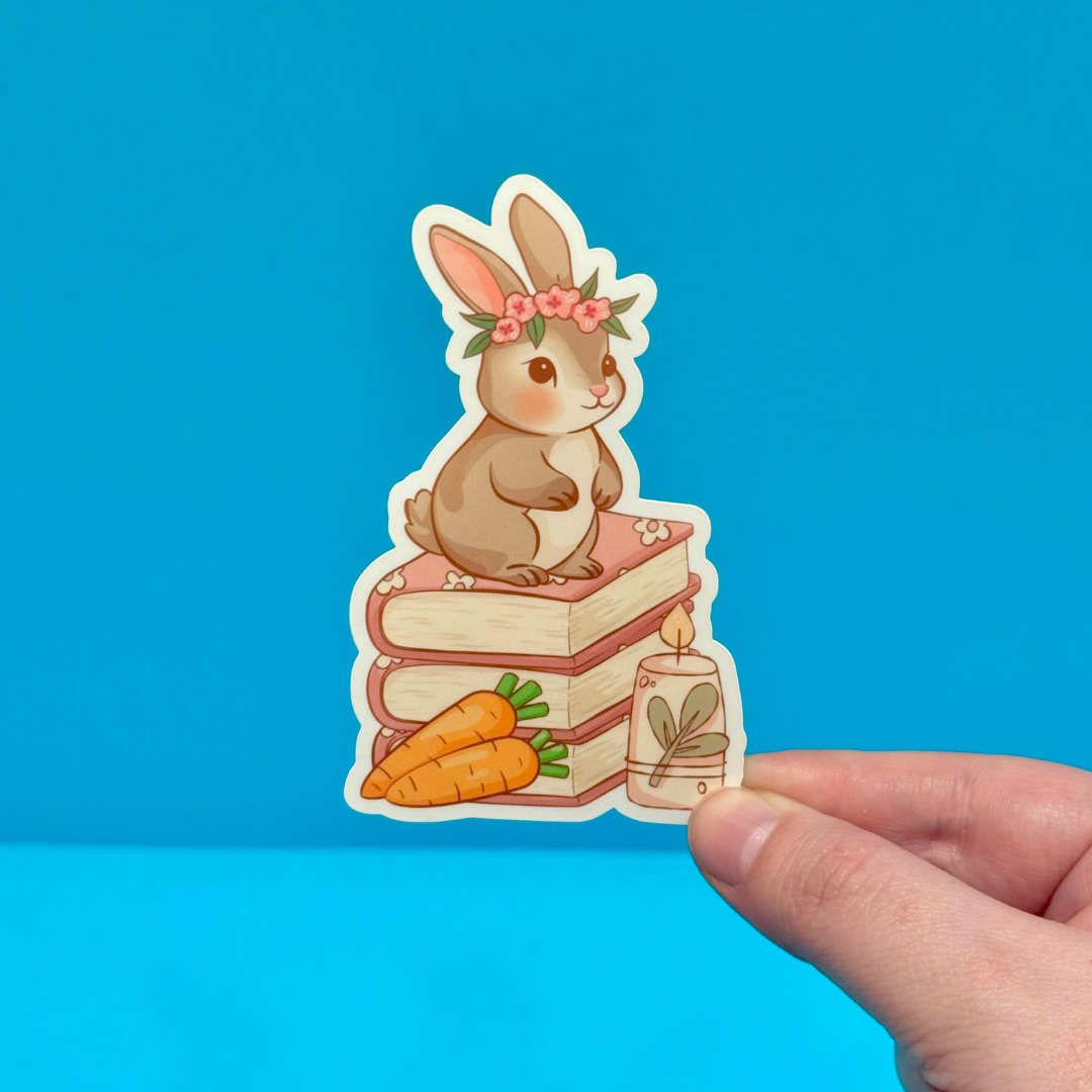 Sticker of a rabbit on books with carrots and a candle, held against a blue background
