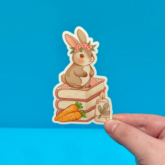 Sticker of a rabbit on books with carrots and a candle, held against a blue background